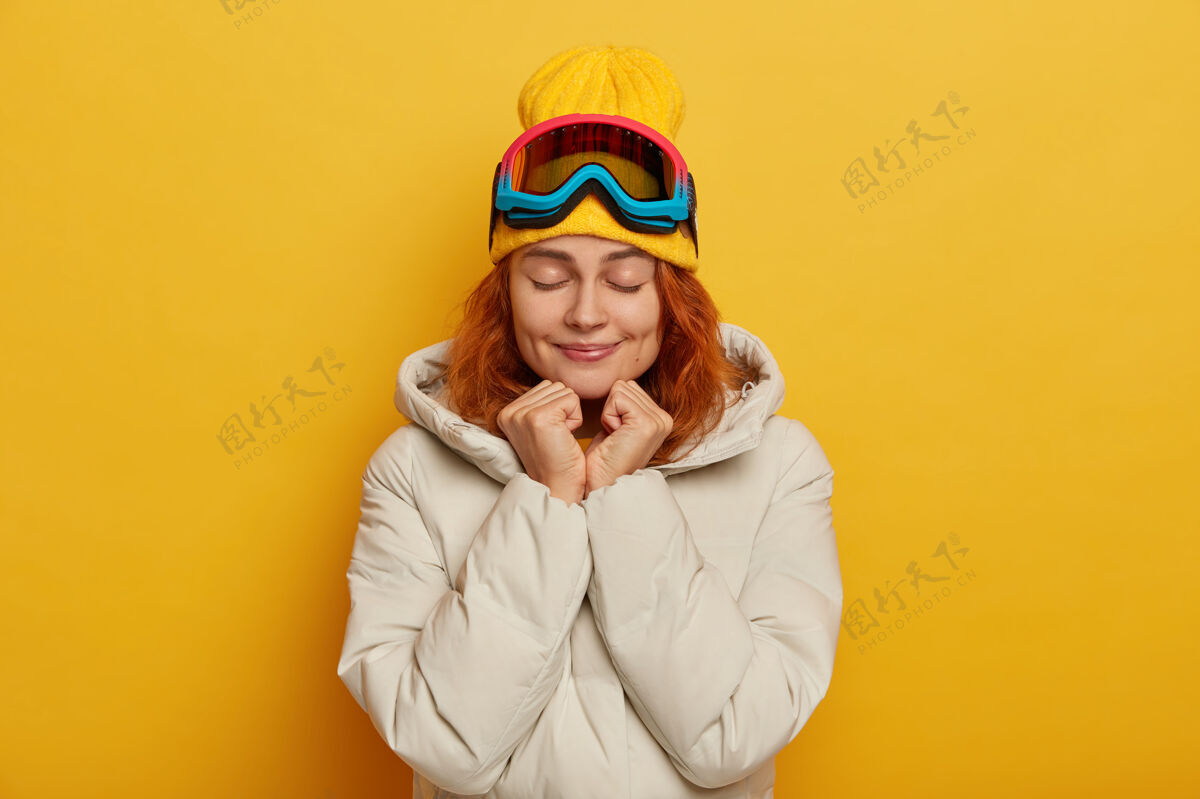 运动滑雪者女孩喜欢业余时间 手放在下巴下面 眼睛闭着 喜欢冬季运动 戴帽子和白色外套 戴着保护性的滑雪面罩 隔离在黄色的墙上护目镜满意酒窝