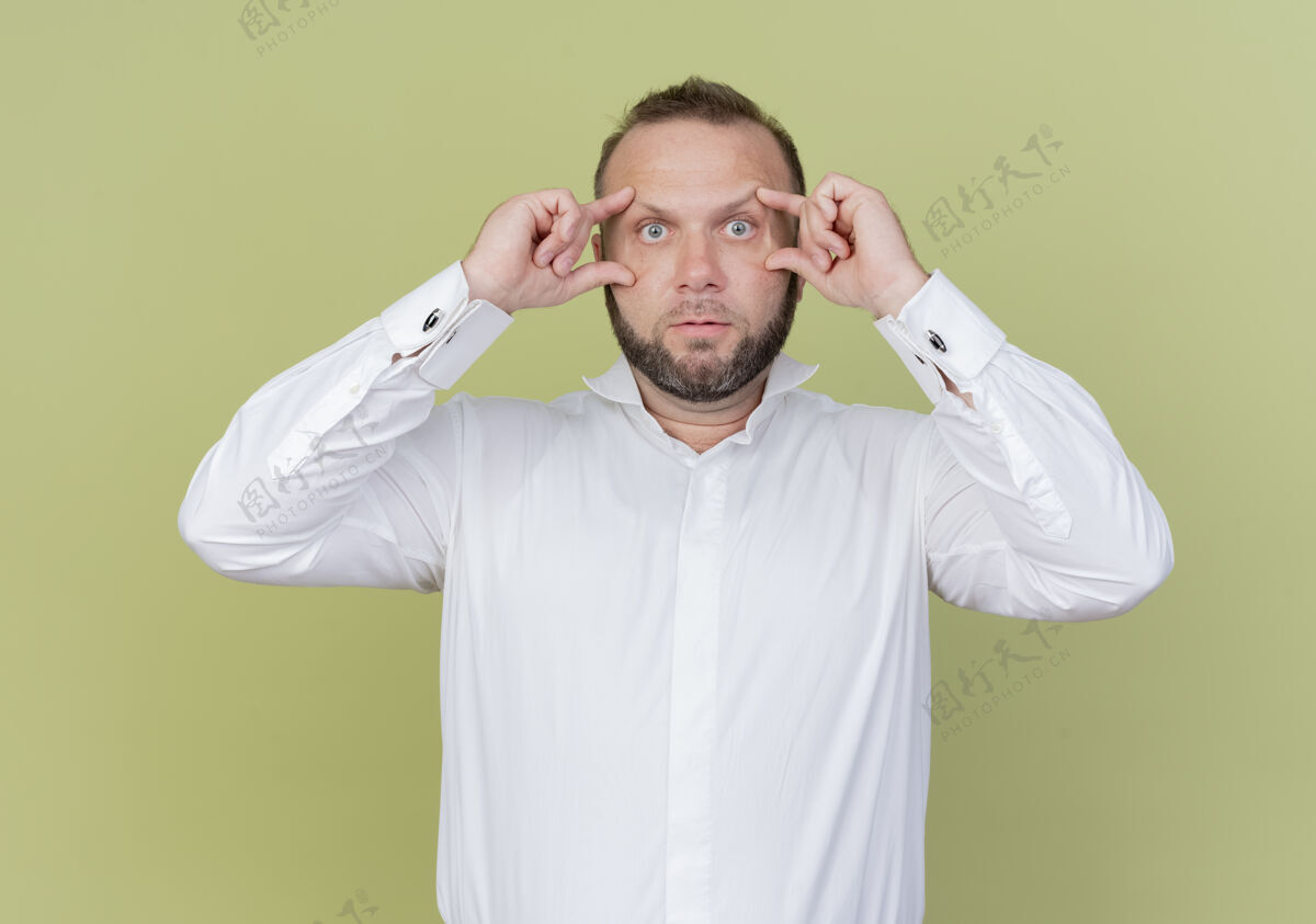 手指一个留着胡子的男人穿着白衬衫睁着眼睛 用手指试图看得更清楚 站在光墙上胡子站穿