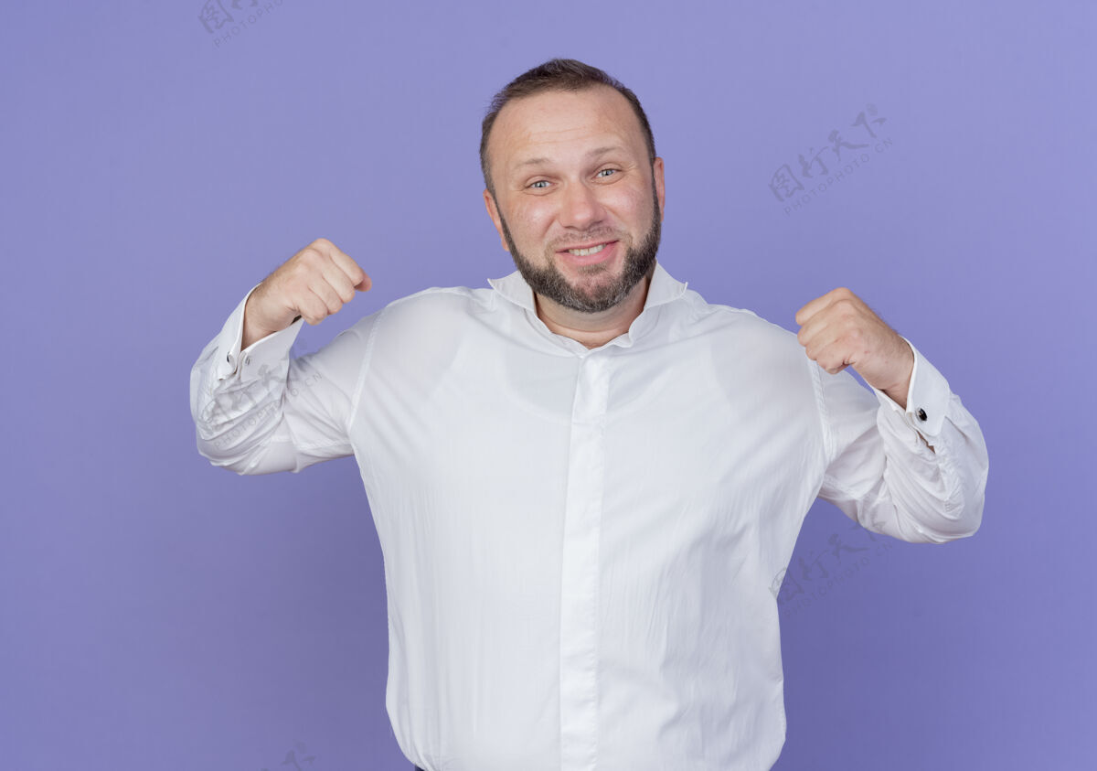 穿一个留着胡子的男人穿着白衬衫 握紧拳头 高兴而兴奋地站在蓝色的墙上为自己的成功而欢呼高兴站立兴奋