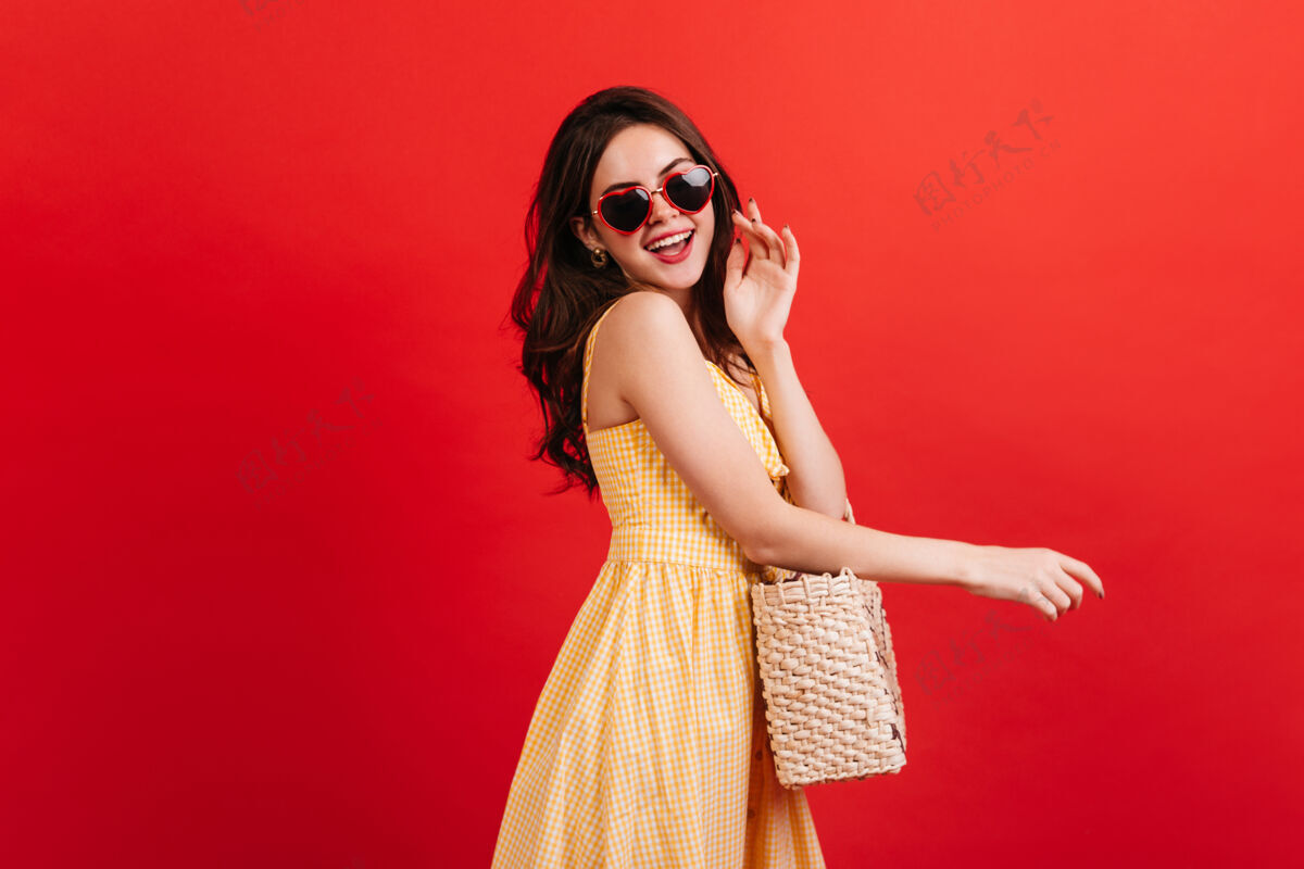 欢呼穿着格子裙的时髦迷人女孩在红墙上欢笑女模特戴着心形眼镜和柳条包的照片自信时尚欢乐