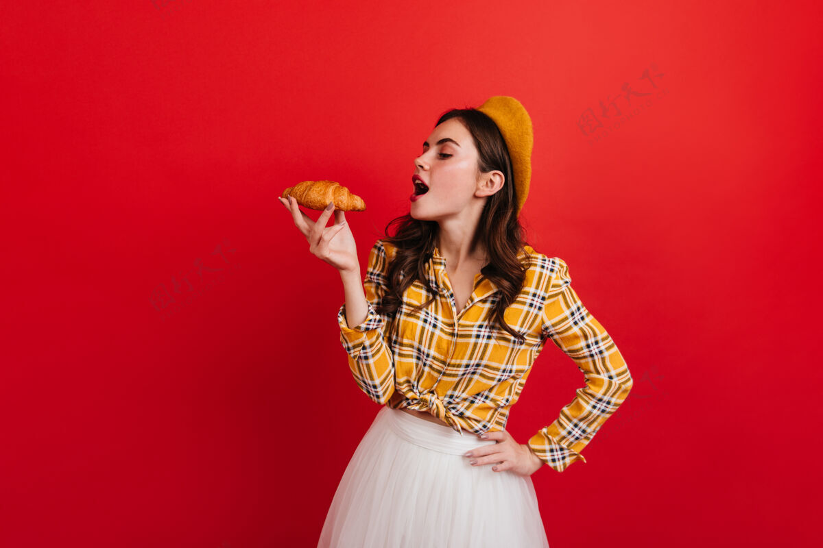 人在红墙上 卷曲的法国女孩吃着酥脆的羊角面包的画像穿着格子衬衫 戴着黄色帽子的黑发女人看着小面包表情贝雷帽波浪发