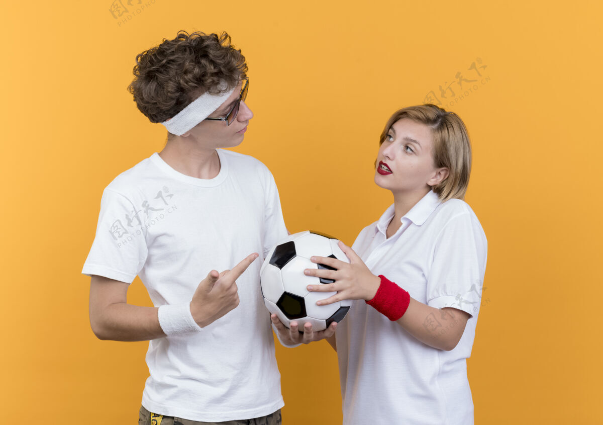 男人年轻的运动型情侣严肃的男人拿着足球看着站在橘色墙上微笑的女友站着运动认真
