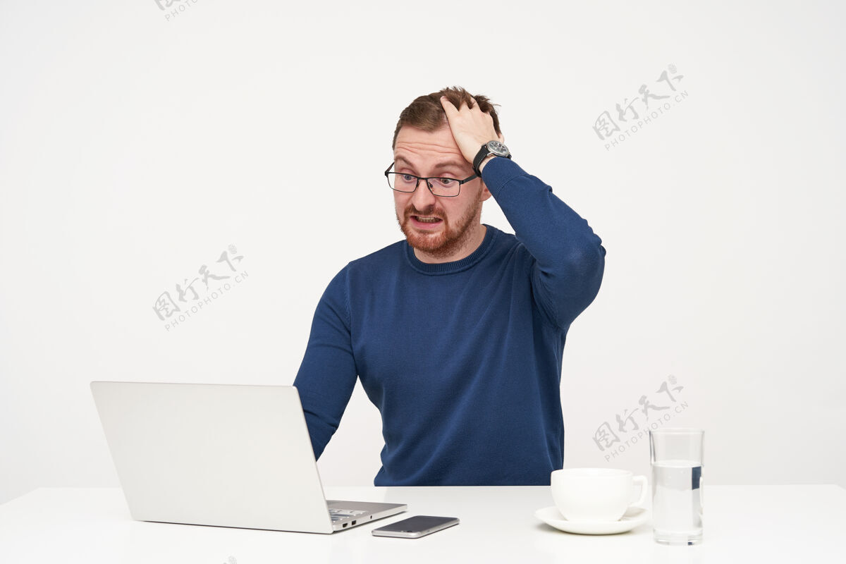 工作激动的年轻帅哥 戴着眼镜 头发乱蓬蓬的 一边惊奇地看着笔记本电脑 一边坐在白色背景上读着意想不到的新闻困惑困惑男士