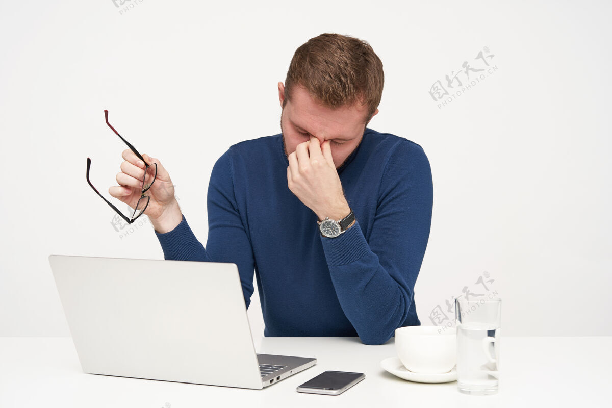 眼镜疲惫不堪的年轻金发男子穿着蓝色毛衣 一边脱下眼镜 一边疲惫地用笔记本电脑工作 隔离在白色背景下笔记本电脑成人水