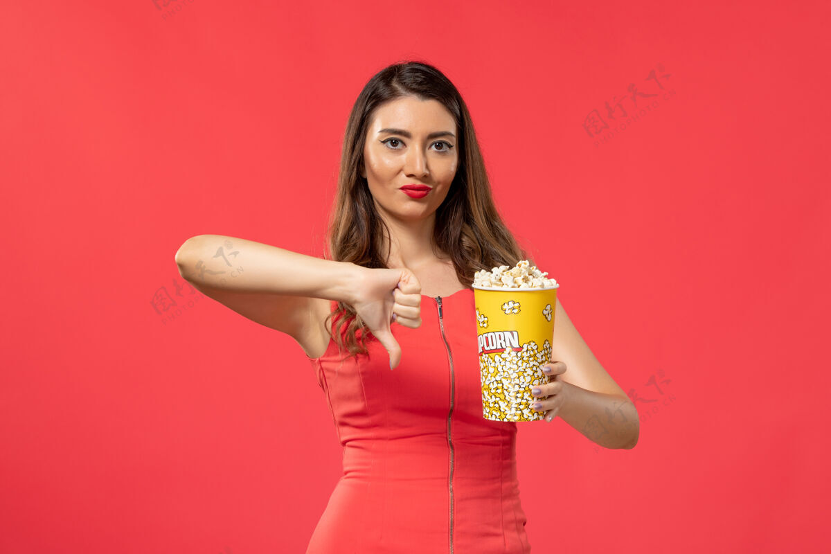 女士正面图身穿红色衬衫的年轻女性手持爆米花在浅红色的表面上前面持有电影