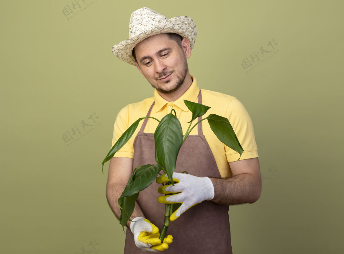 连身衣年轻的园丁 穿着连体衣 戴着帽子 戴着工作手套 手里拿着一株植物 面带微笑地看着它年轻人看抱