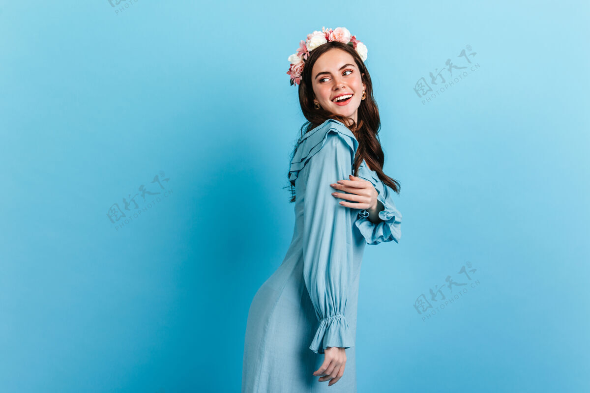 自信温柔的女孩带着雪白的微笑环顾四周戴着花冠的模特和丝质连衣裙摆在蓝色的墙上服装肖像手