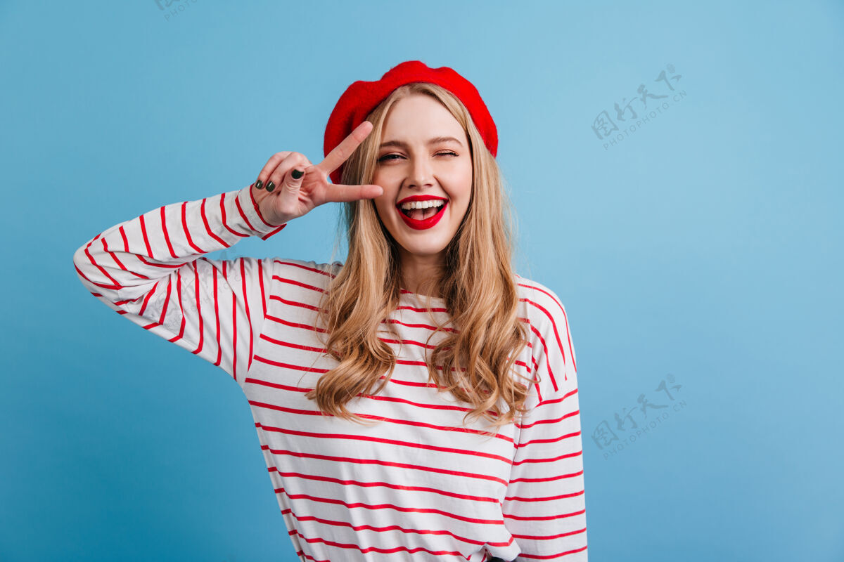 时尚美丽的金发女孩在条纹衬衫显示和平标志前视图的法国女士笑着摆在蓝色的墙上欢呼和平化妆