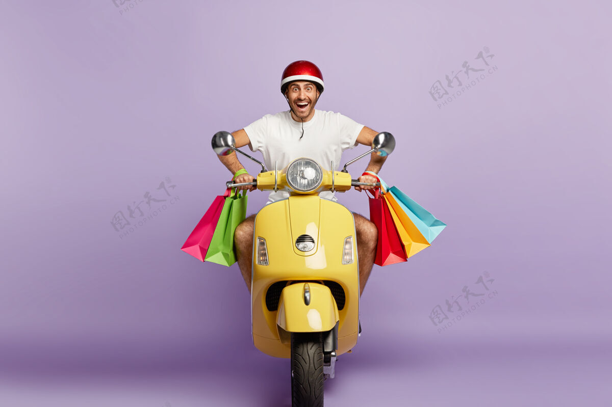 购物带着头盔和购物袋的快乐微笑的家伙开着黄色的滑板车T恤五颜六色年轻