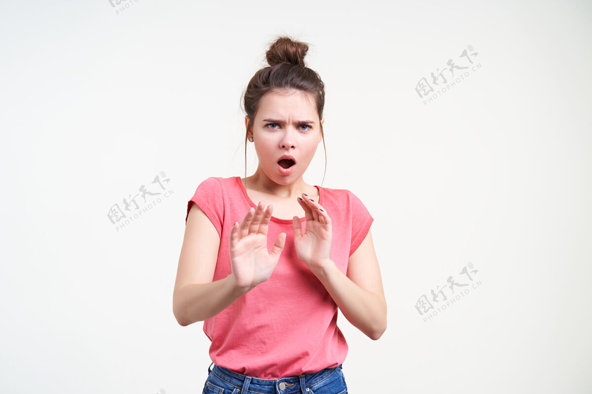T恤摄影棚拍摄的一张照片 害怕的棕色头发的年轻女性惊恐地看着相机 张开嘴 举起双手做着停止的手势 站在白色的背景上害怕粉色鬼脸
