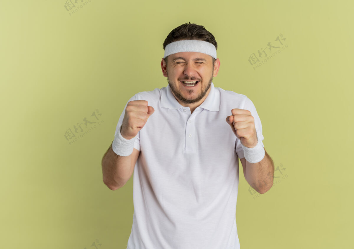 健身身穿白衬衫 头箍紧握拳头的年轻健身男子站在橄榄色背景下为自己的成功感到高兴和兴奋握紧成功白色