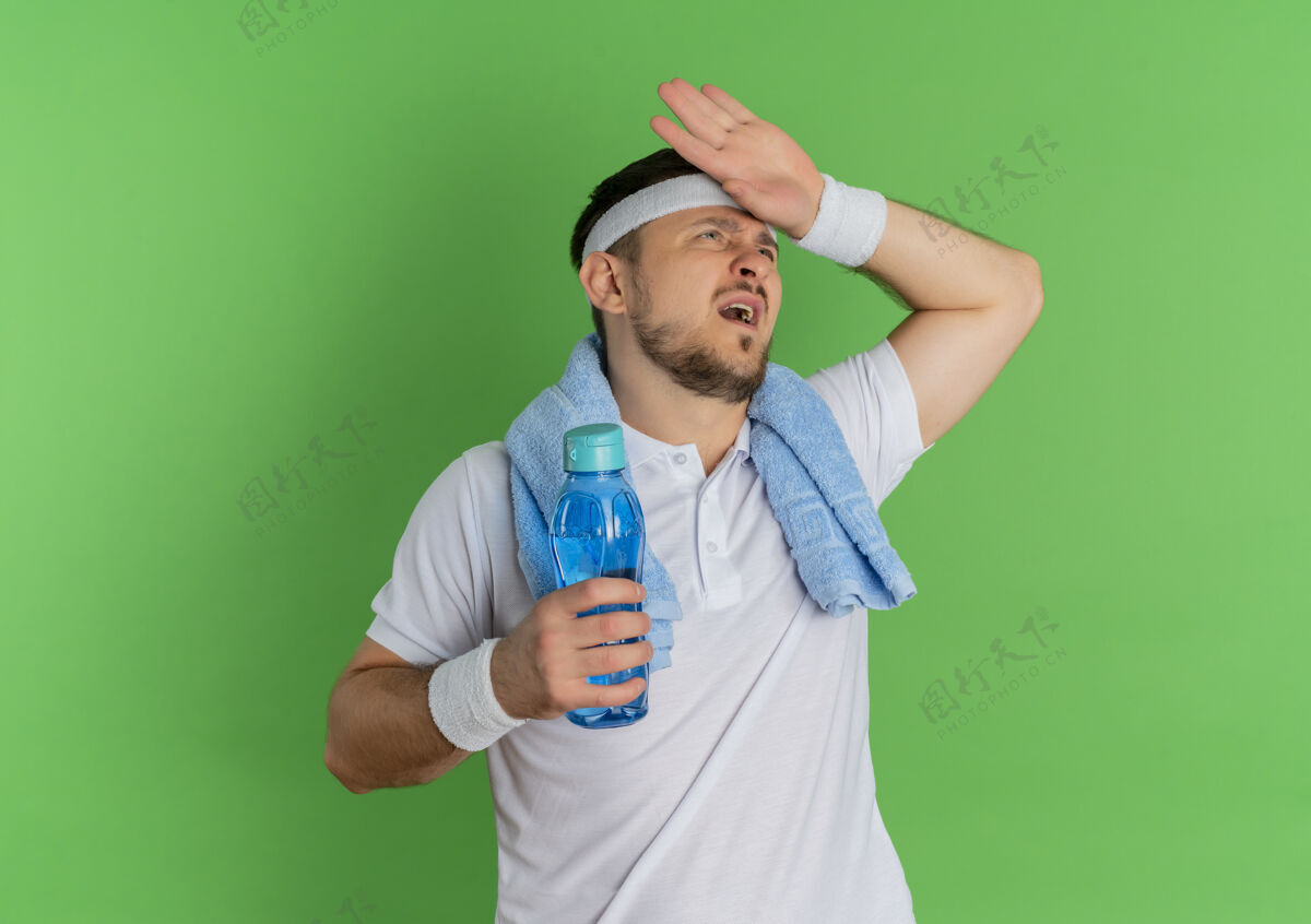 瓶子身穿白衬衫 头戴头巾 脖子上围着毛巾 手拿一瓶水的年轻健身男子站在绿色背景上 累得筋疲力尽白色抱着衬衫