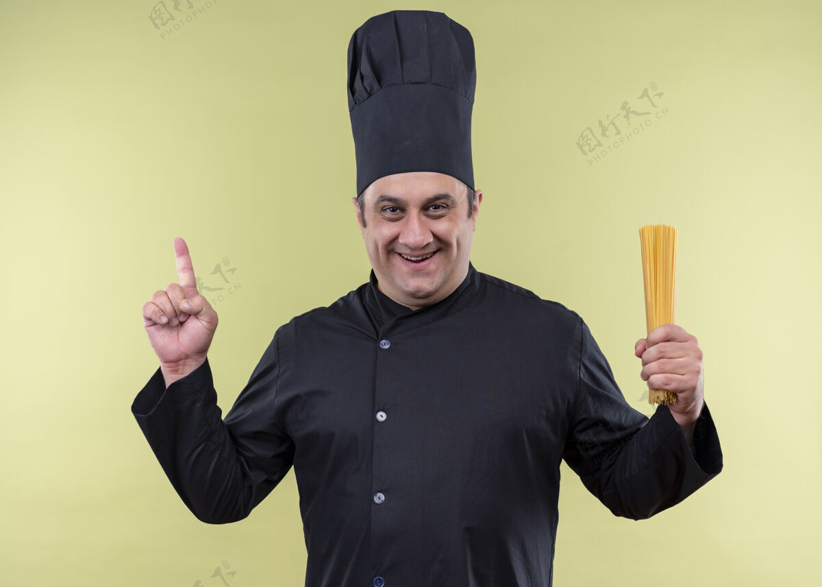帽子男厨师身穿黑色制服 头戴厨师帽 手拿一排意大利面 手指朝上看着摄像机 站在绿色背景下 满脸笑容指着站着微笑