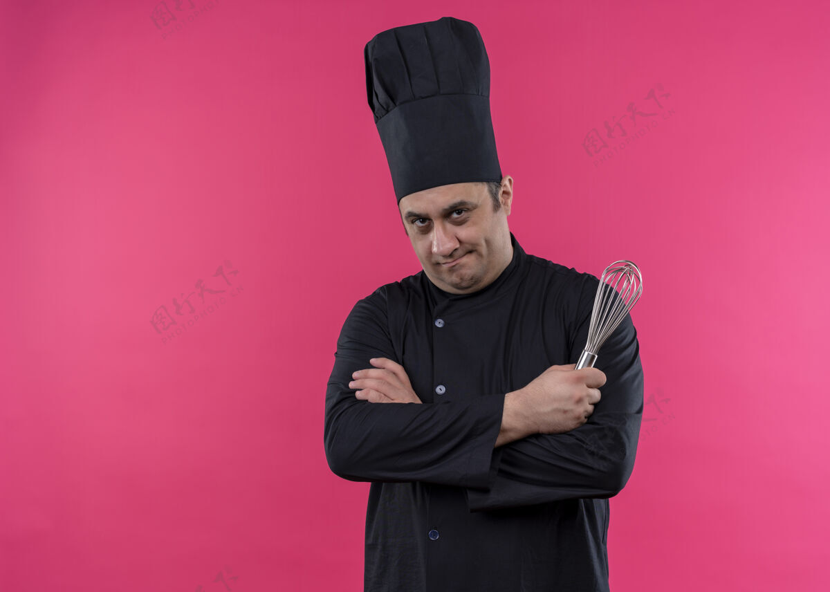 帽子男厨师身穿黑色制服 头戴厨师帽 手拿花冠 表情严肃 双臂交叉站在粉色背景上 看着镜头粉色脸抱着