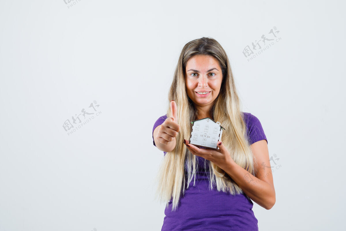显示年轻的女士拿着房子模型 展示拇指在紫罗兰色的t恤衫 看起来很高兴前视图时尚时尚女性