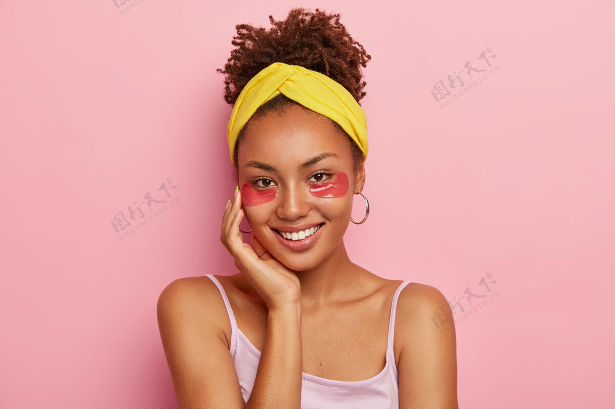 人微笑的非裔美国妇女的画像 戴着眼罩 减轻浮肿 眼袋 摸脸颊 把卷发梳成发髻 戴着头带 耳环 微笑愉快女性牙刷年轻