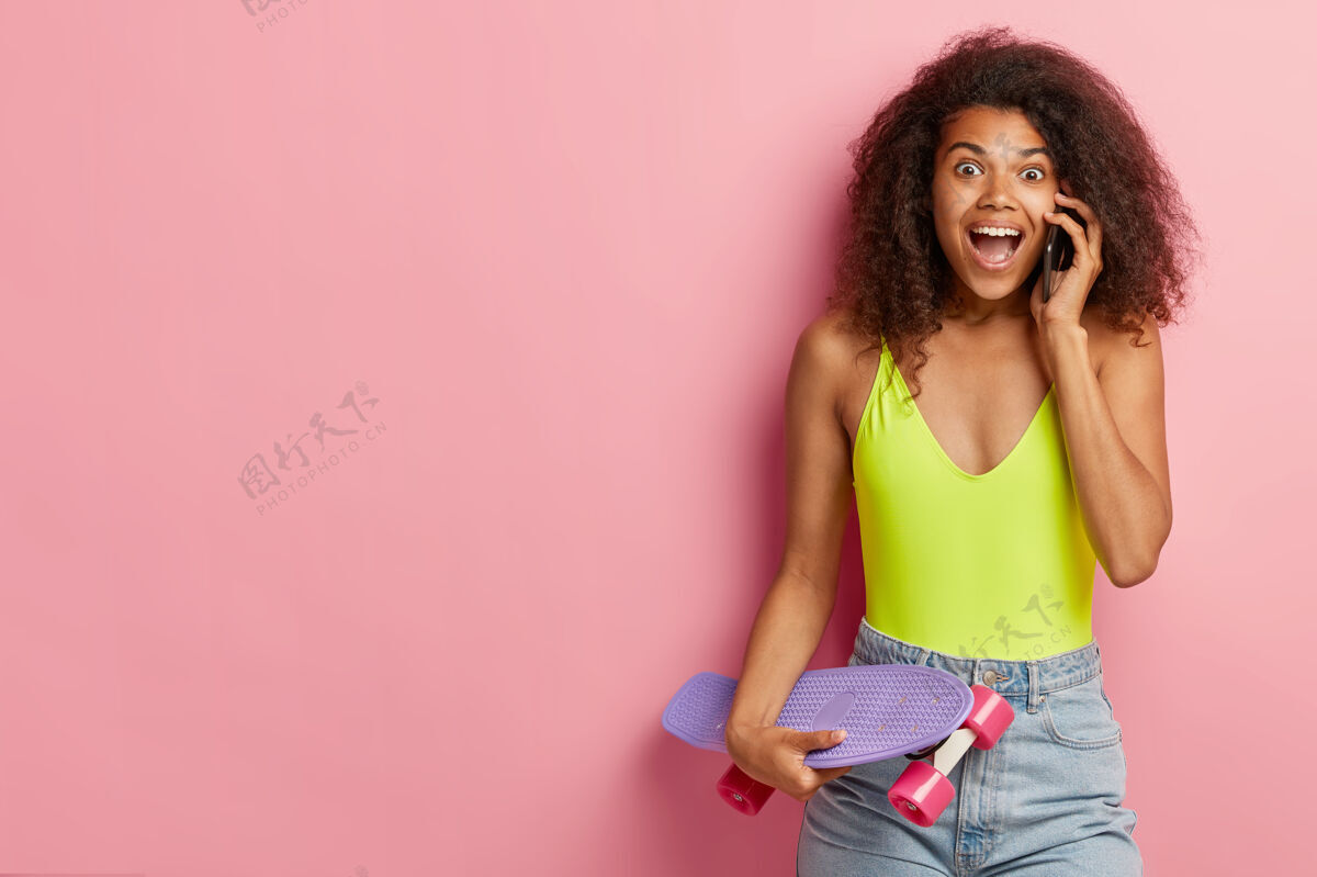 爱好情绪化的黑皮肤女人通过手机说话 告诉朋友新闻 穿着时髦的夏装 拿着长板 靠着粉色的墙站着 把空间放在一边生活方式的概念粉色乐观移动
