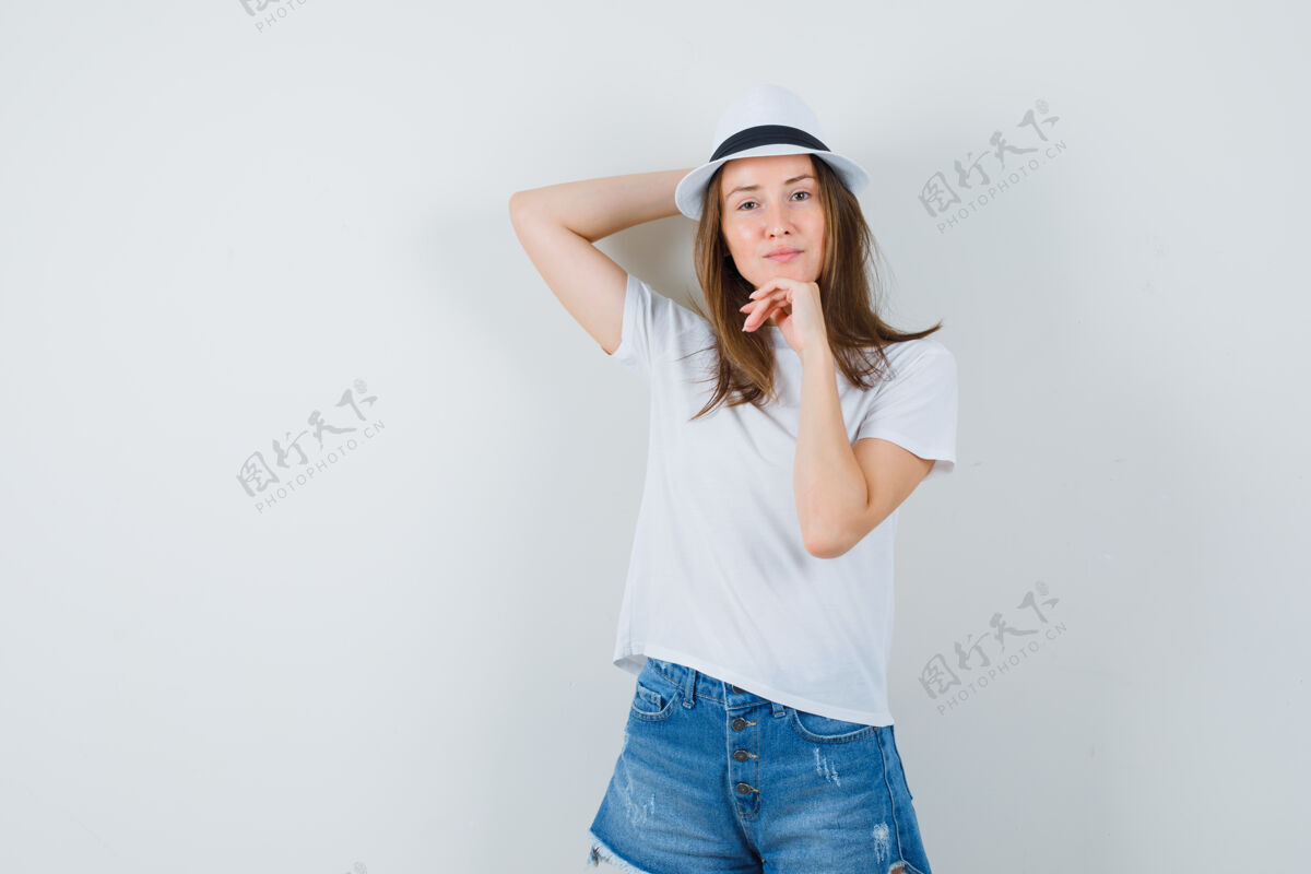 时尚穿着白色t恤 短裤 帽子的年轻女子站着摆姿势 看起来很讨人喜欢欢呼女性模特