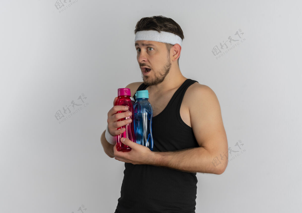 头带戴着头巾拿着几瓶水的年轻健身男子站在白色背景下 看上去很困惑水困惑健身