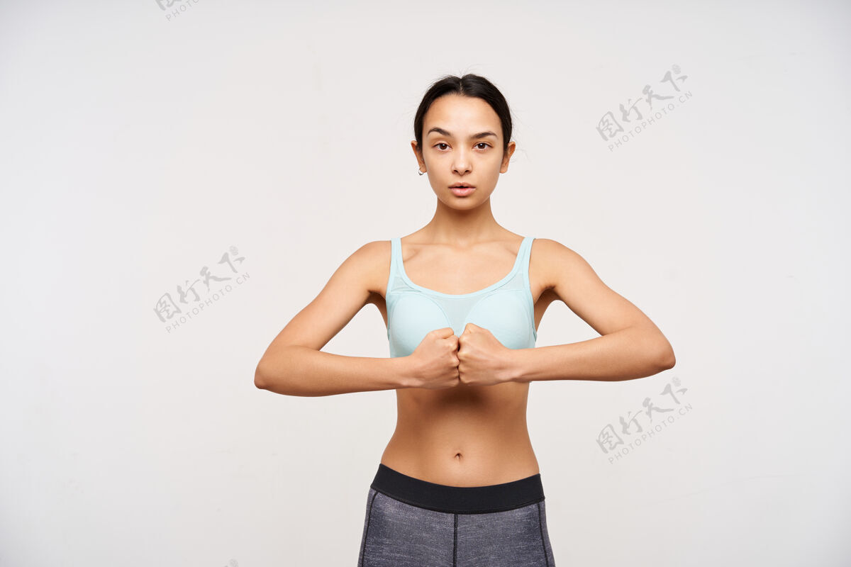 二头肌摄影棚拍摄了一位年轻的棕色头发的运动女士 她把拳头叠在一起 同时威胁地看着前面 穿着运动服站在白色的墙上强壮运动装女性