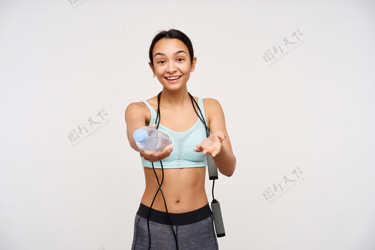 女士活跃的年轻积极的黑发女性微笑愉快地拿着一瓶水 脖子上戴着跳绳站在白色的墙壁上站立高兴锻炼