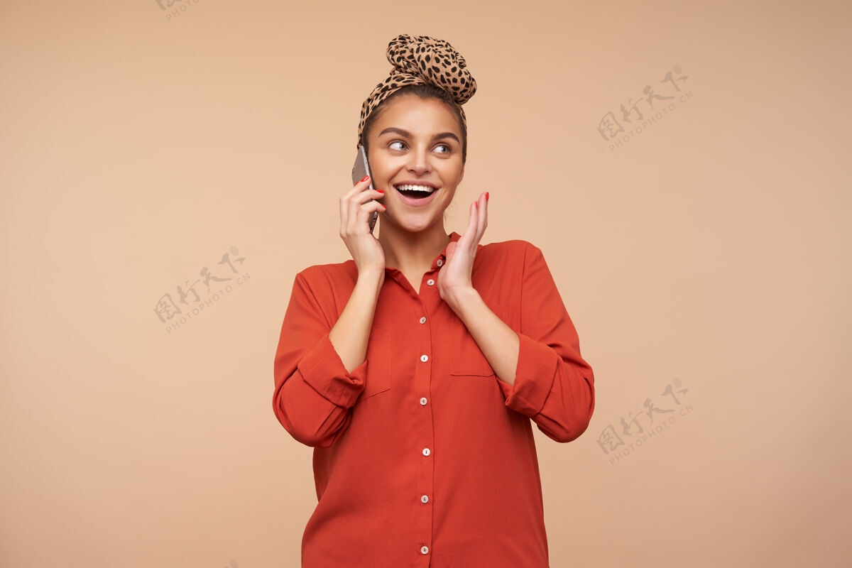 心情快乐的年轻迷人的棕色头发的女人 自然的妆容微笑愉快 同时有愉快的电话交谈 隔离在米色的墙壁发髻豹子配件