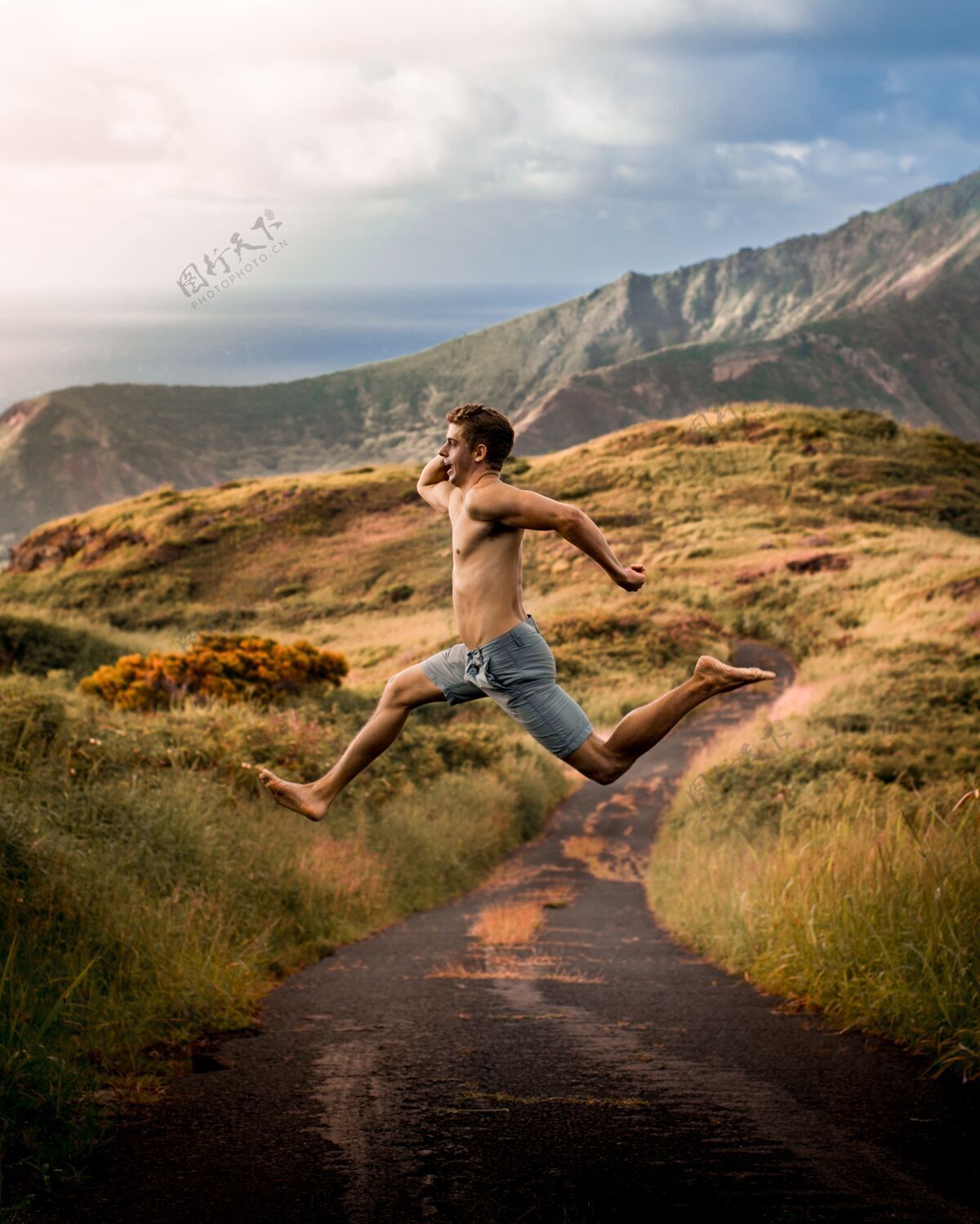 运动在阳光和多云的天空下 年轻的雄性在群山环绕的田野里跳跃太阳草地男孩
