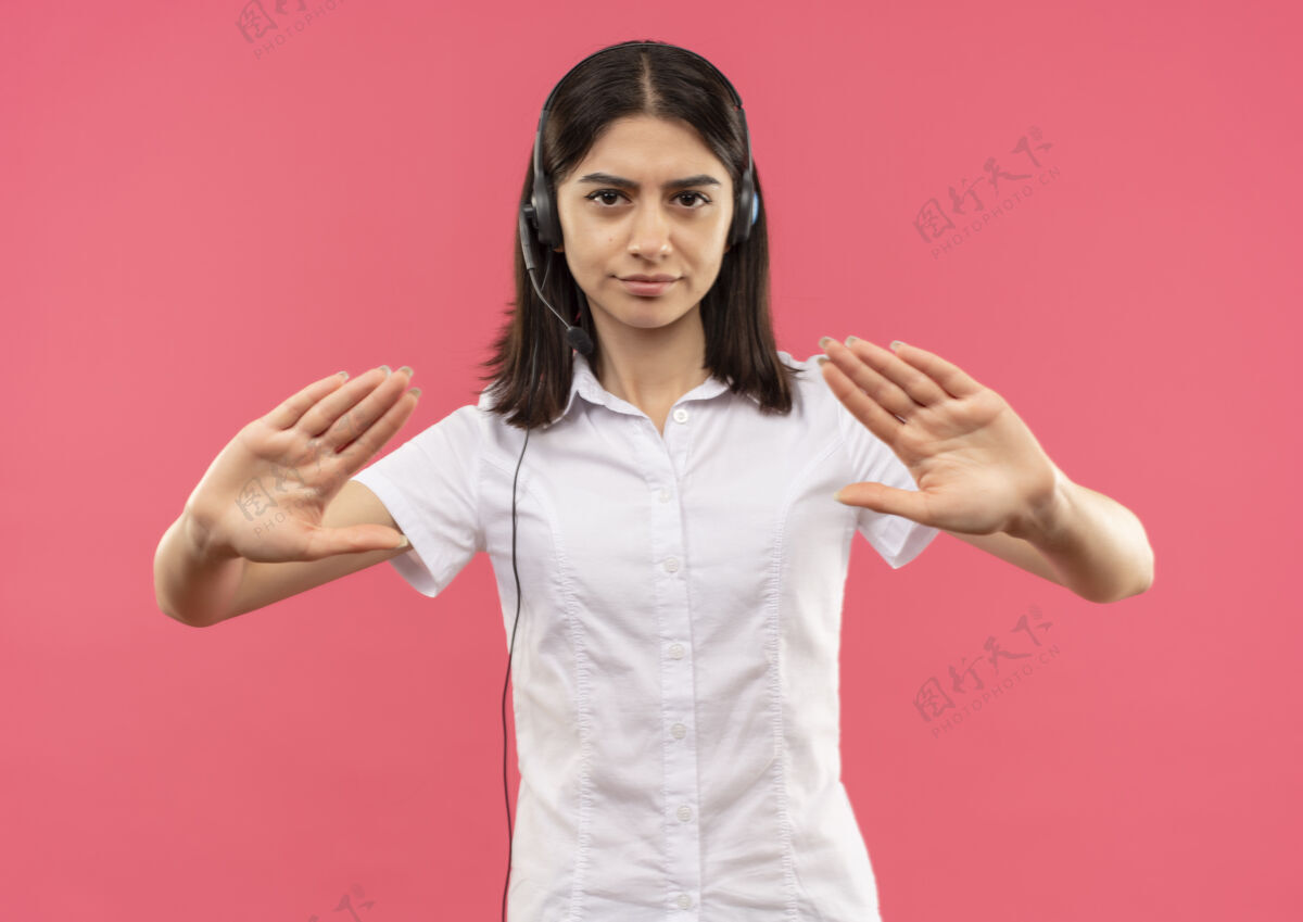 年轻人穿着白衬衫戴着耳机的年轻女孩 手朝前看 严肃的脸站在粉红色的墙上 做着停车标志公民麦克风衬衫