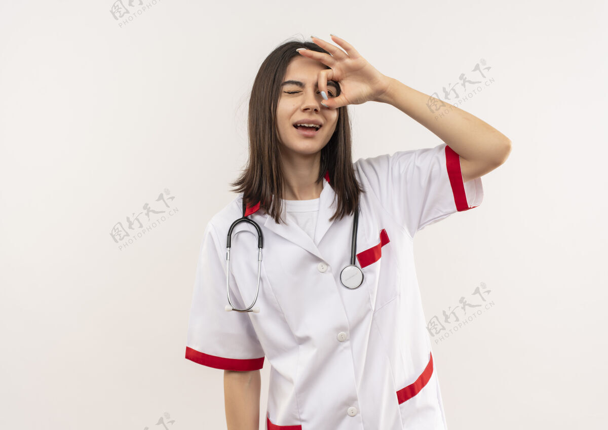 周围年轻的女医生穿着白大褂 脖子上戴着听诊器 做着“ok”的手势 站在白墙上看着这首歌女通过站