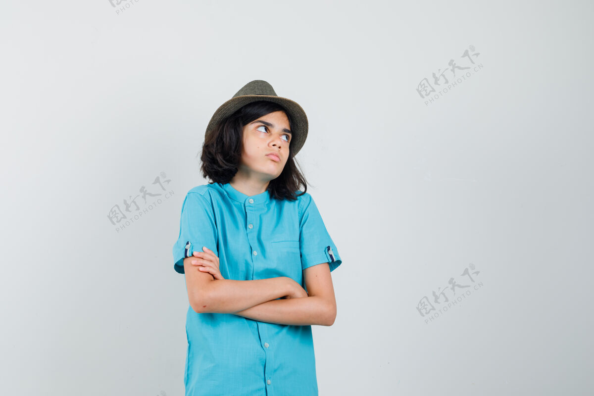 手臂穿蓝色衬衫 戴着帽子的年轻女士双手交叉站着 一边看着一边 看起来很复杂积极喜悦肖像