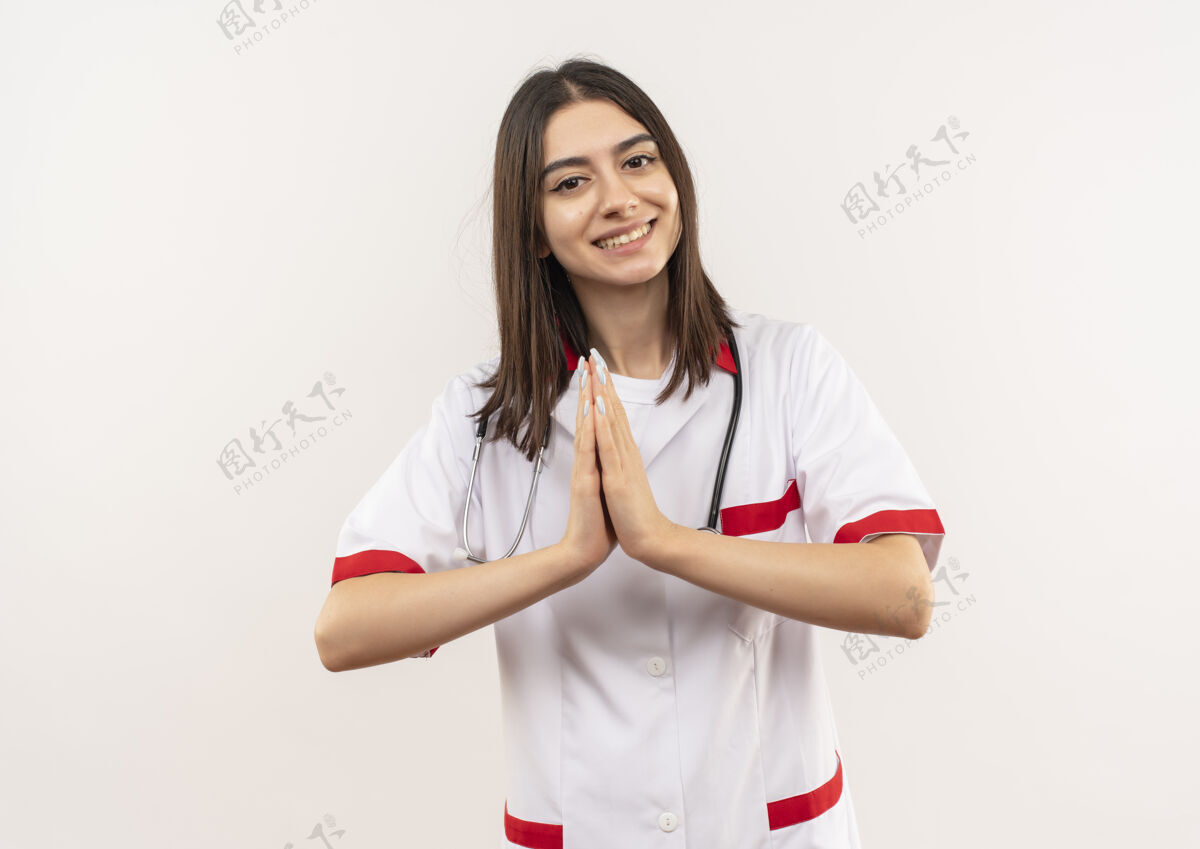 姿势年轻的女医生穿着白大褂 脖子上戴着听诊器 双手握在一起 站在白墙上表示感谢站着姿势围着