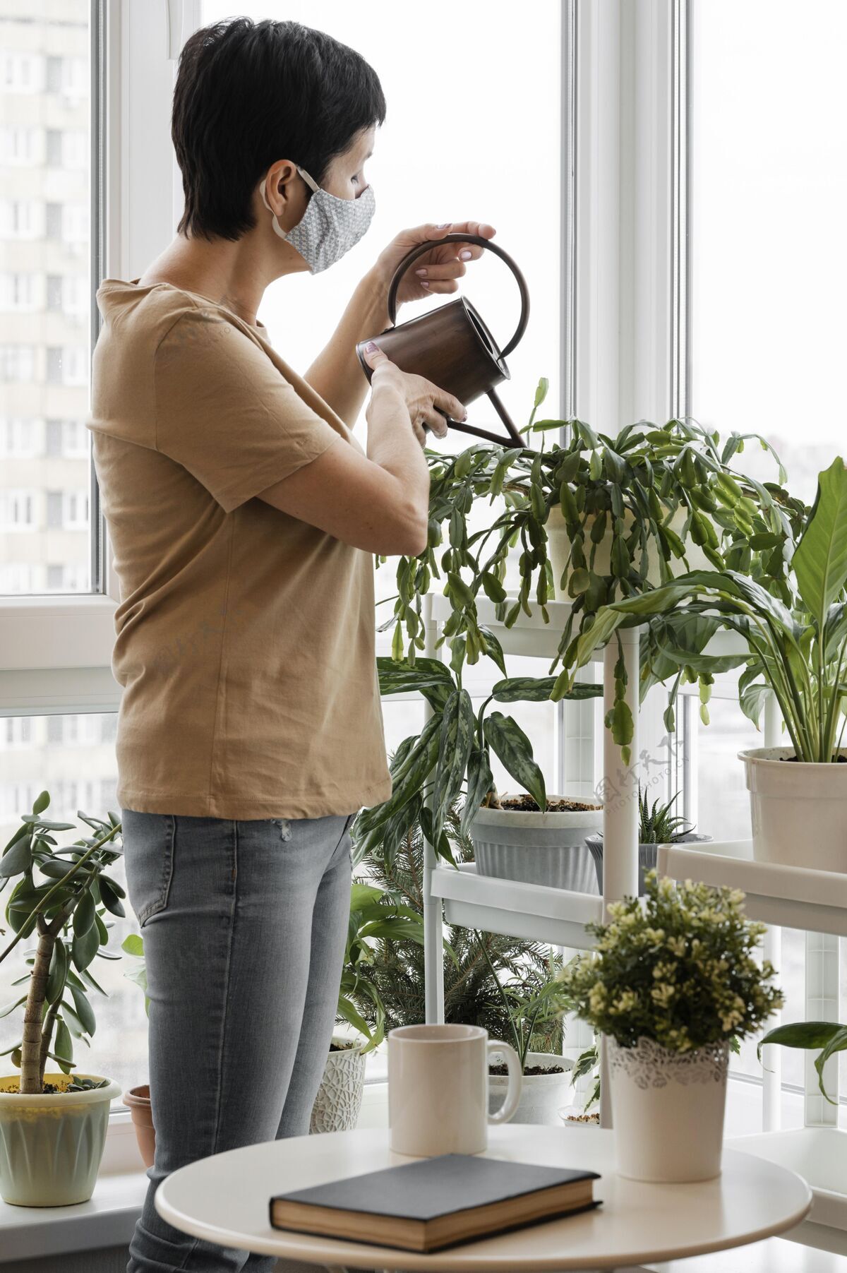 草药戴着口罩在花盆里浇灌室内植物的女人的侧视图家庭室内花卉