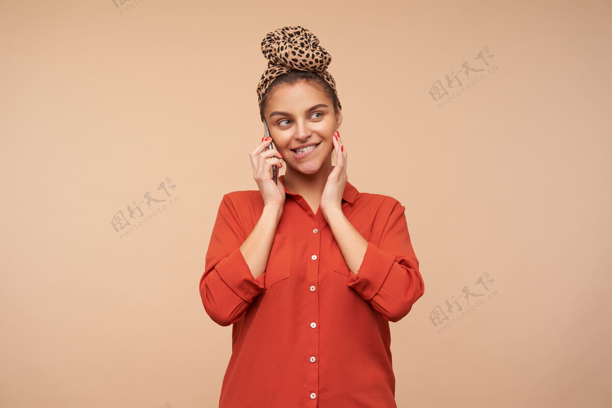 女性年轻可爱的棕色头发的女士 自然化妆 一边打电话一边用手抚摸着脸 咬着屁股 在米色的墙上摆姿势手机站着发型