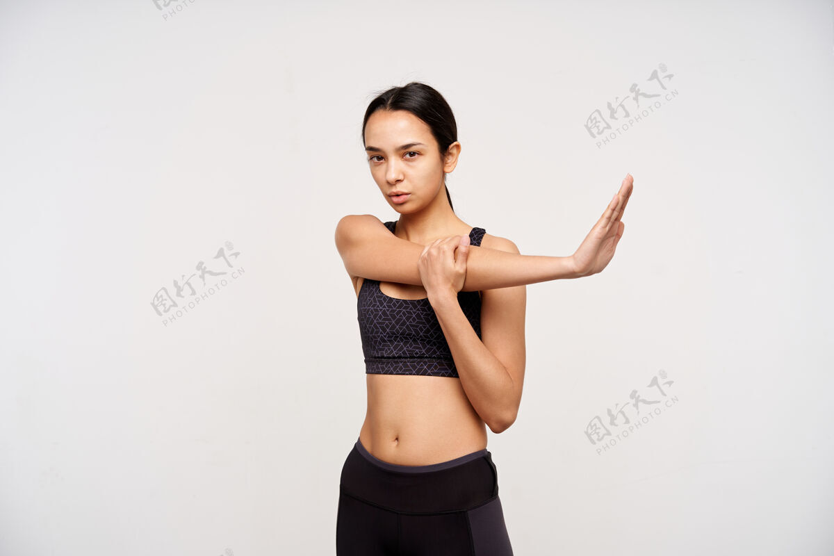 头发摄影棚拍摄的年轻运动黑发女子穿着运动服伸展双臂 认真地看着前面 隔着白色的墙壁 穿着运动服自然爱手