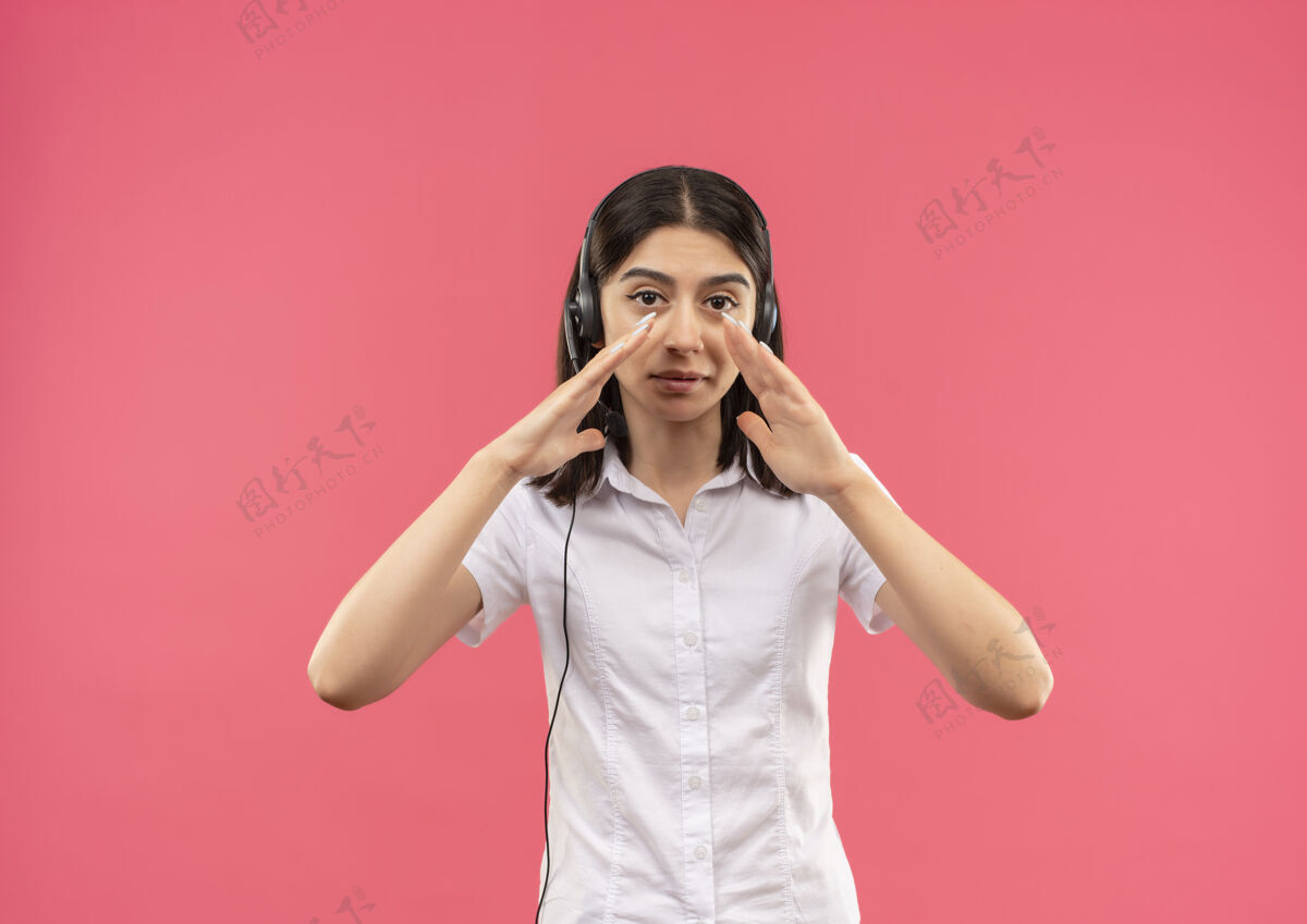 嘴穿着白衬衫戴着耳机的年轻女孩 站在粉红色的墙上 用手靠近嘴巴喊着或叫着什么人站立呼叫有人