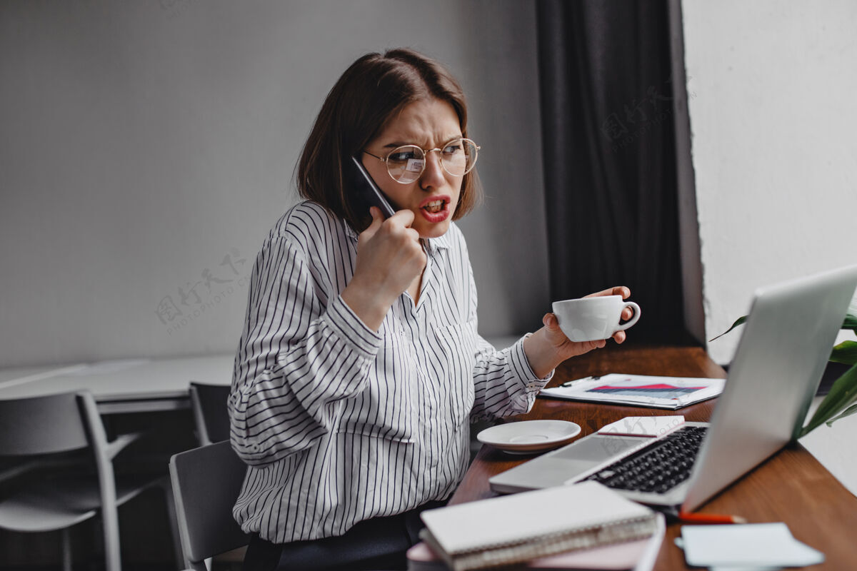 商务女性愤怒的商人和下属通电话不满意的女员工穿着白衬衫拿着白杯子坐在桌旁拿着笔记本电脑电话笔记本电脑成年人