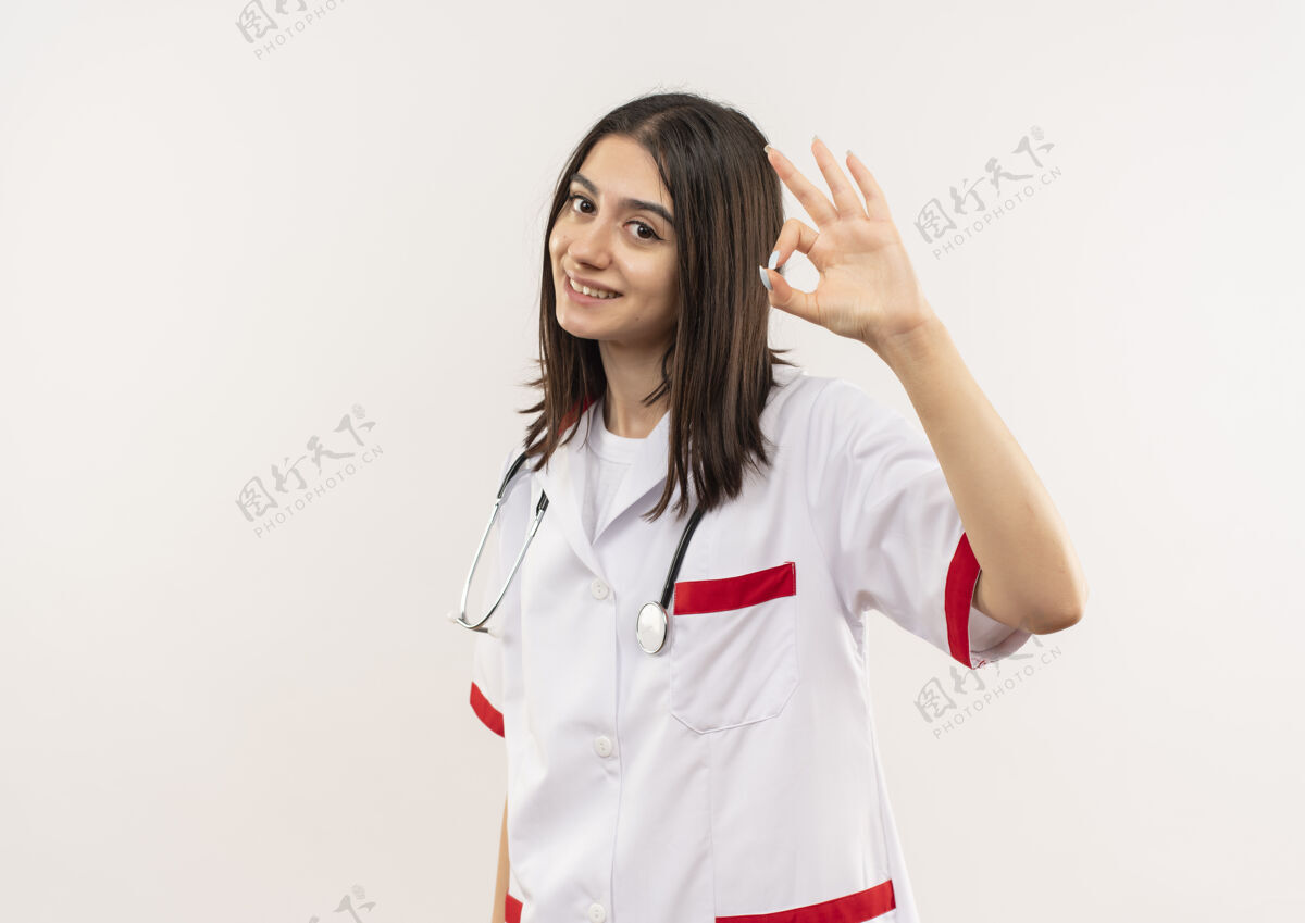 周围年轻的女医生穿着白大褂 脖子上戴着听诊器 面带微笑 友好地站在白色的墙上 显示“ok”标志看微笑女人