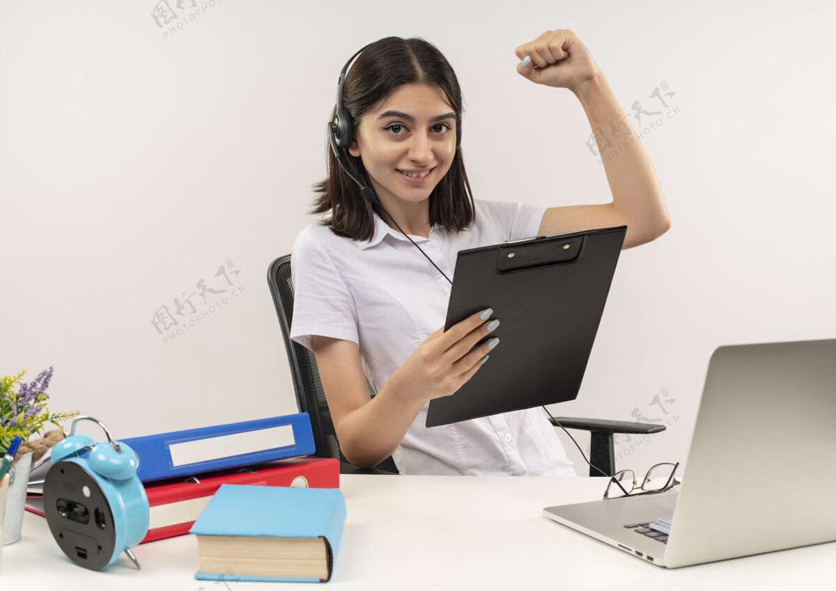坐着穿着白衬衫戴着耳机的年轻女孩 拿着写字板握紧拳头快乐而积极地坐在桌子旁 把文件夹和笔记本电脑放在白墙上快乐女孩笔记本电脑