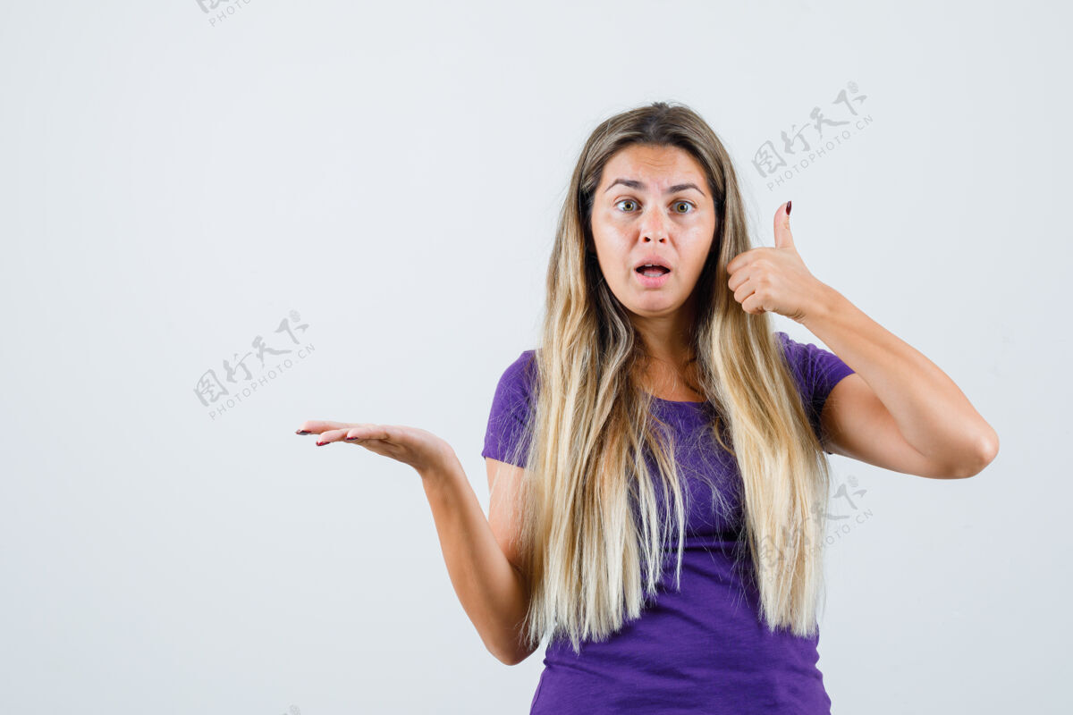 女孩在紫罗兰色t恤的正面视图中 金发女孩向上伸出拇指 同时将手掌放在一边时尚年轻肖像