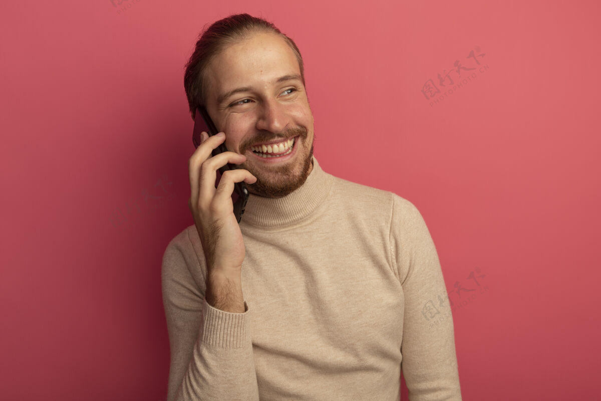 同时站在粉红色的墙上 穿着米色高领毛衣的年轻帅哥一边高声微笑一边讲着手机微笑男人姿势