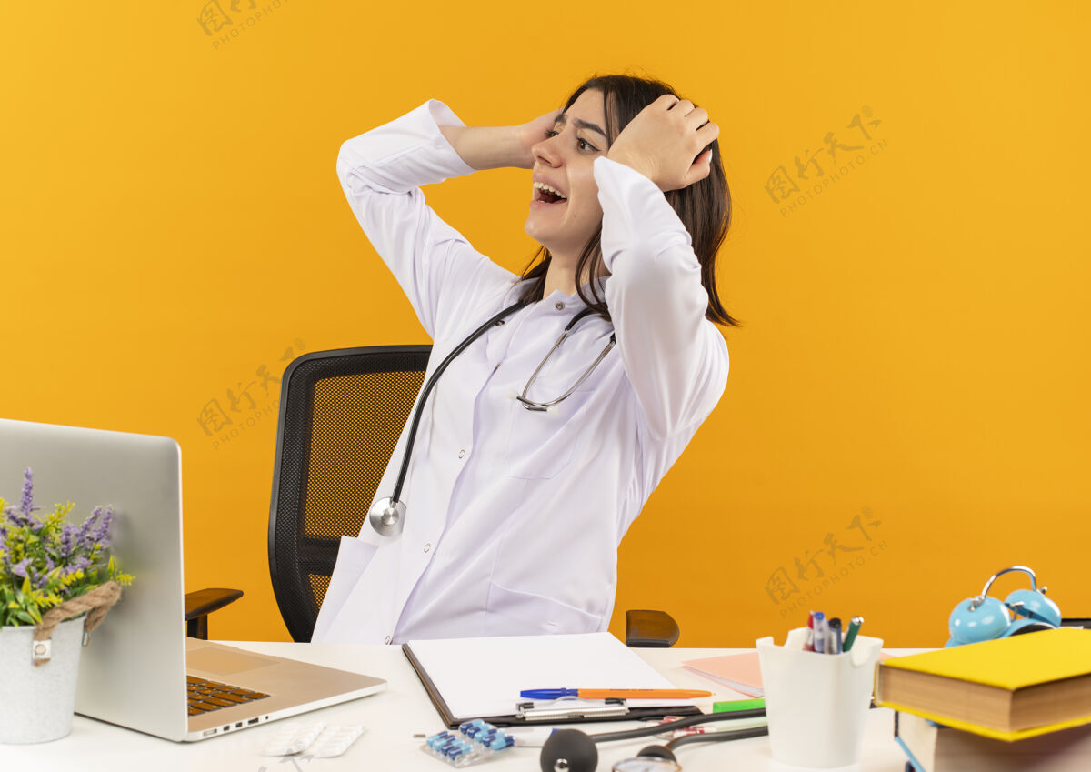 拿着年轻的女医生穿着白大褂 手持听诊器 面带微笑地把头放在一边 坐在桌边 手提电脑和文件放在橙色的墙上听诊器外套人