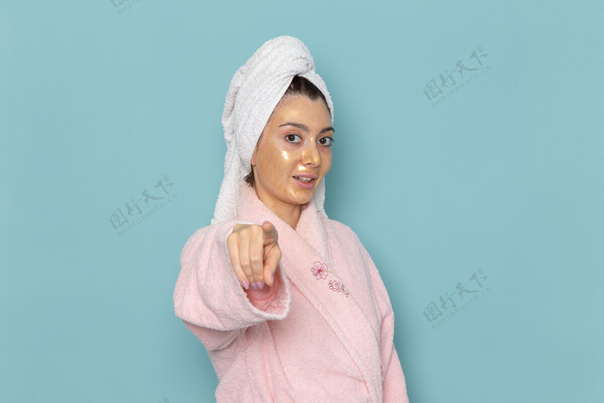 肖像正面图身着粉色浴袍的年轻女性在蓝色墙壁上淋浴后的美丽水浴霜自护淋浴浴室浴缸女帽子