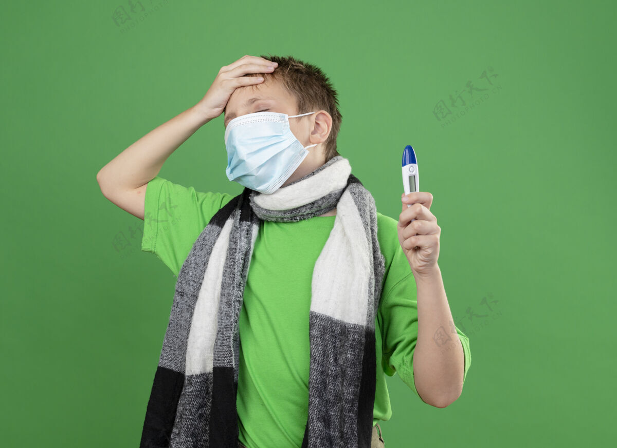 围巾生病的小男孩穿着绿色t恤 脖子上围着暖和的围巾 戴着护面面具 手里拿着温度计 手放在头上 站在绿色的墙上 神情困惑 非常焦虑手面具非常
