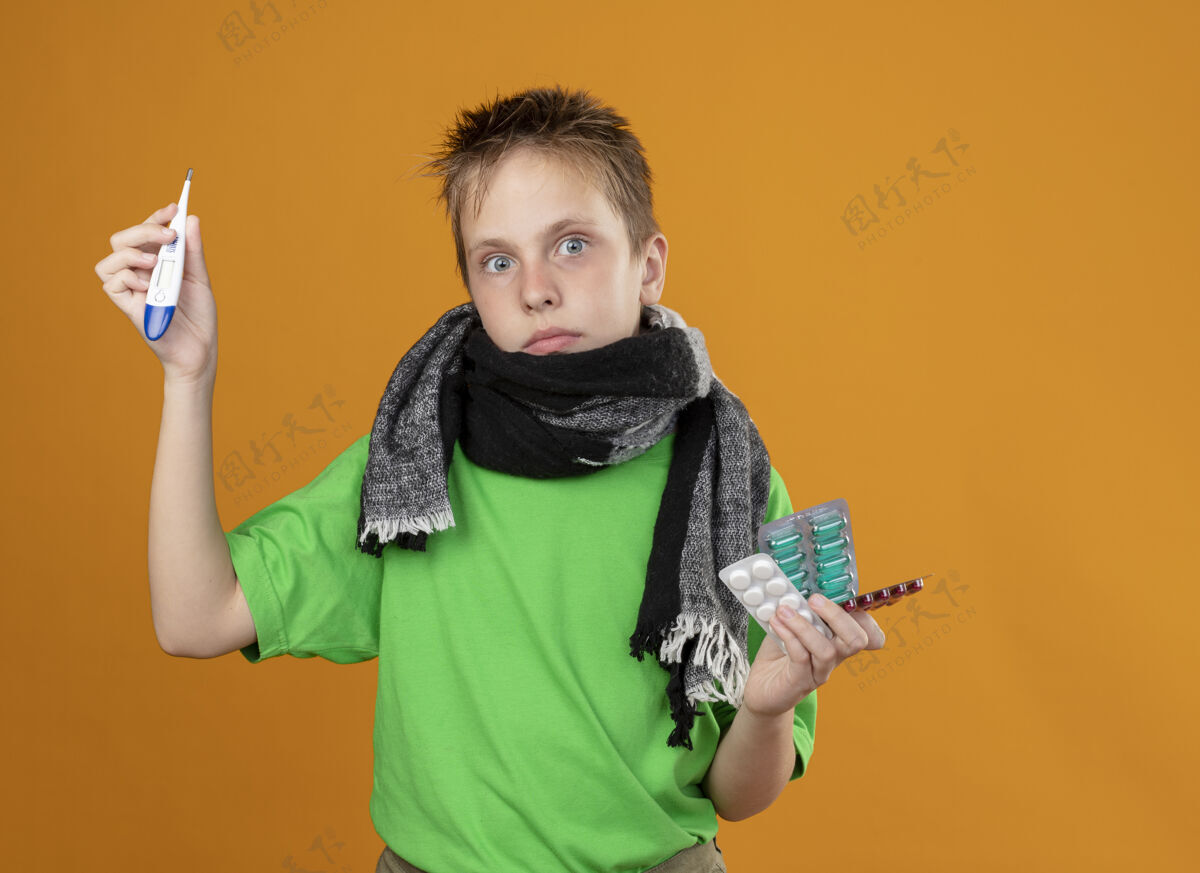 围巾生病的小男孩穿着绿色t恤 脖子上围着暖和的围巾 手里拿着药片 站在橙色的墙上 看上去很困惑男孩脖子看着