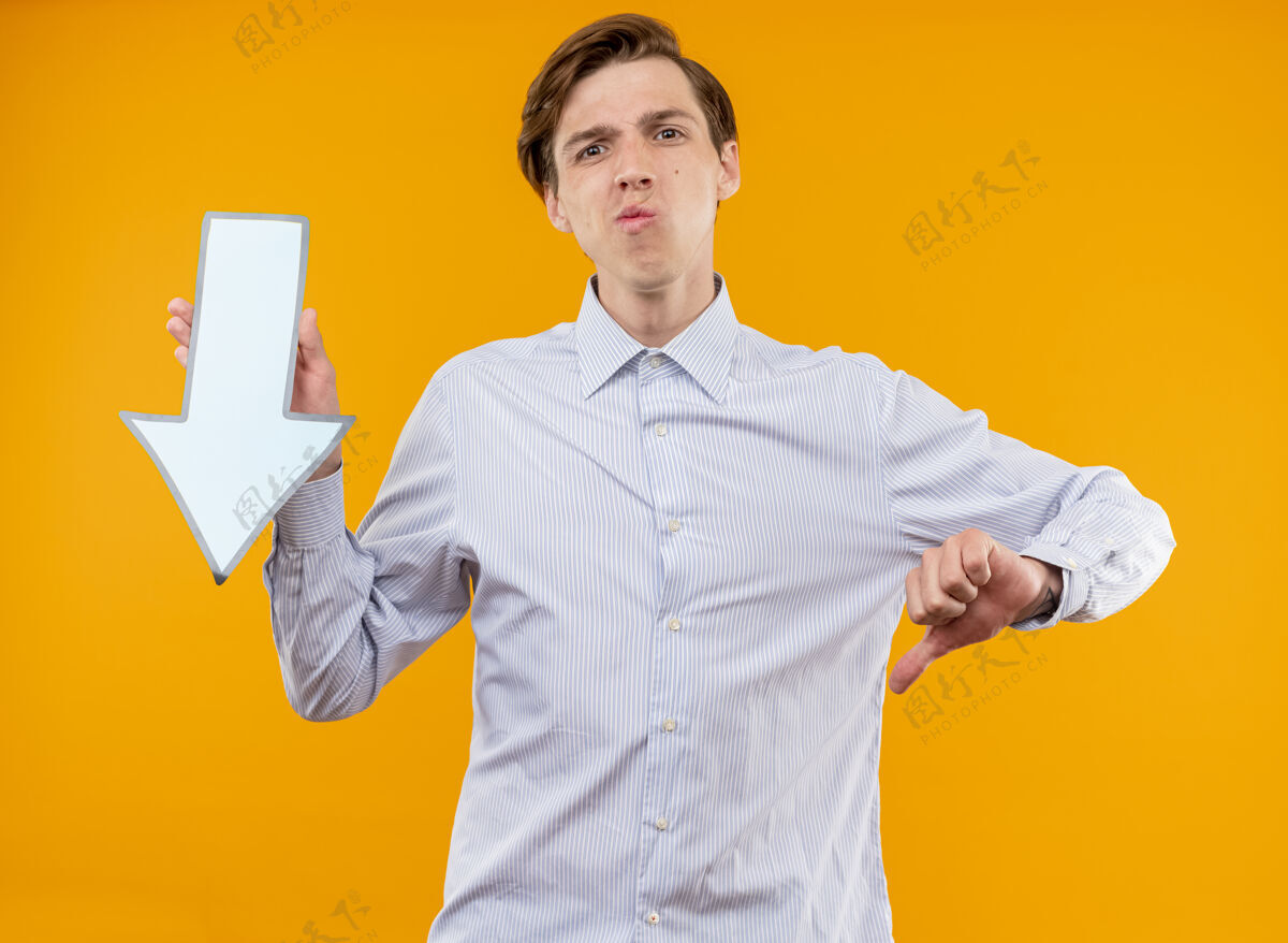 显示身穿白衬衫的年轻人举着蓝色箭头 不高兴地伸出大拇指站在橙色的墙上拇指看站