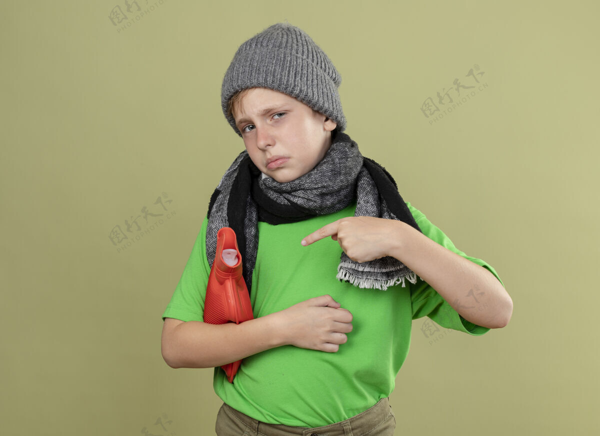 男孩生病的小男孩穿着绿色t恤 戴着暖和的围巾和帽子 感觉不舒服 拿着水瓶取暖 手指指着它站在明亮的墙上围巾少温暖
