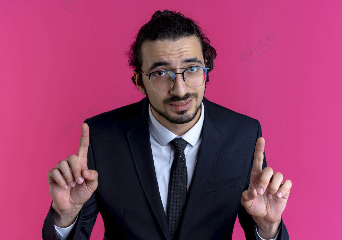 看一个穿着黑色西装戴着眼镜的商人望着前面 食指站在粉红色的墙上手指男人优雅