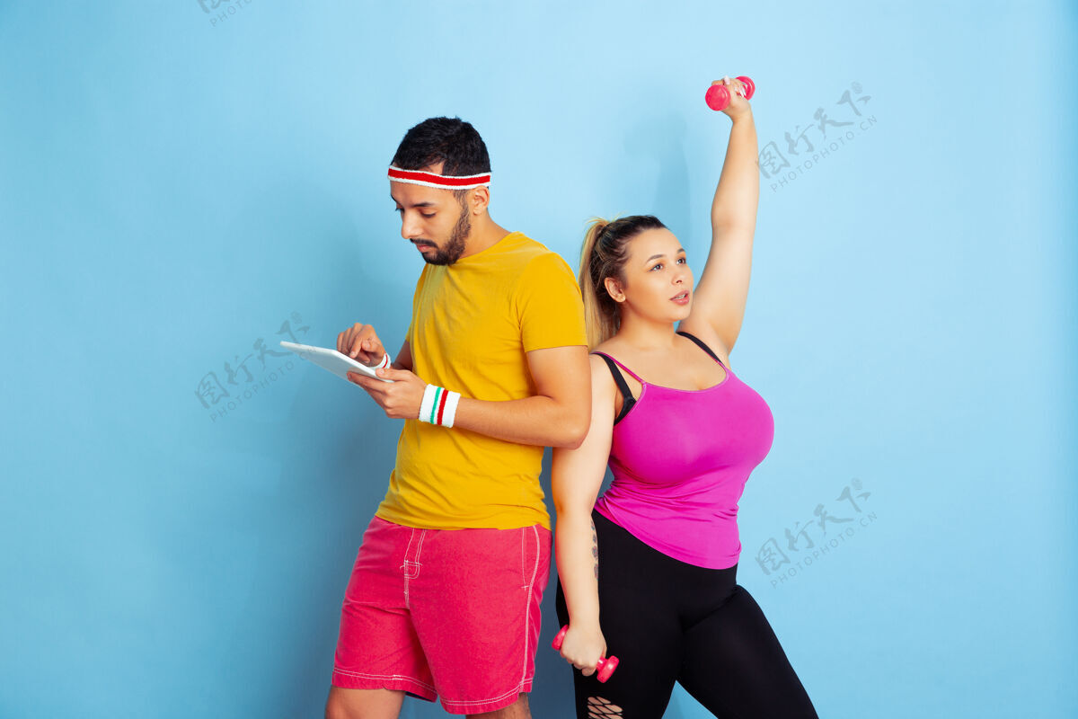 举重年轻漂亮的白人夫妇穿着鲜艳的衣服在蓝色背景上训练运动概念 人类情感 表达 健康的生活方式 关系 家庭她在训练 他在用平板电脑护理肖像体重