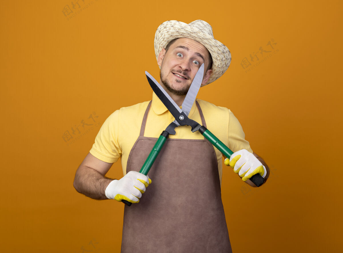 人有趣的年轻园丁穿着连体衣 戴着帽子 手持树篱剪 站在橙色的墙上 面带微笑地看着前面人树篱剪子