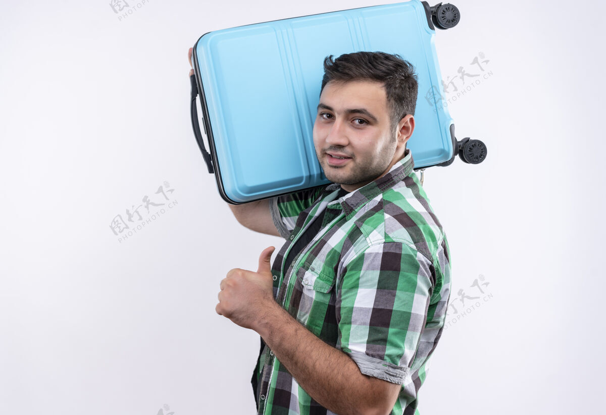 假期穿着格子衬衫的年轻旅行家手拿手提箱 竖起大拇指 微笑着自信地站在白墙上旅行假期公民